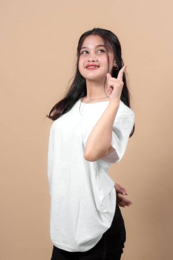 Genç Asyalı kadın gülümsüyor ve işaret parmağıyla yukarıya doğru işaret ediyor. İlham verici ve düşünceli görünüyor. Beyaz bir tişört giyiyor. Stüdyoda bej arka planda izole edilmiş..