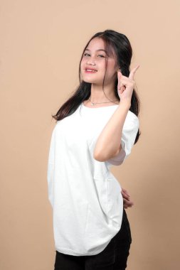 Kendinden emin, gülümseyen ve işaret parmağıyla işaret eden genç bir Asyalı kadın. Tarafsız bej arka planda beyaz tişört giyiyor. Reklam veya markalaşma konsepti için stüdyo çekimi.