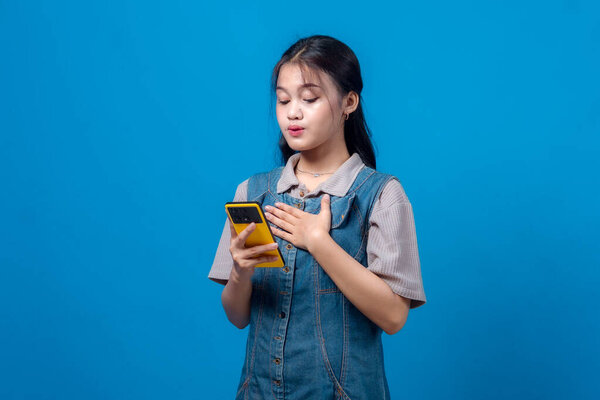 Young Asian woman holding a yellow smartphone with one hand on her chest, expressing surprise or gratitude, standing against a plain blue background, looking emotional.