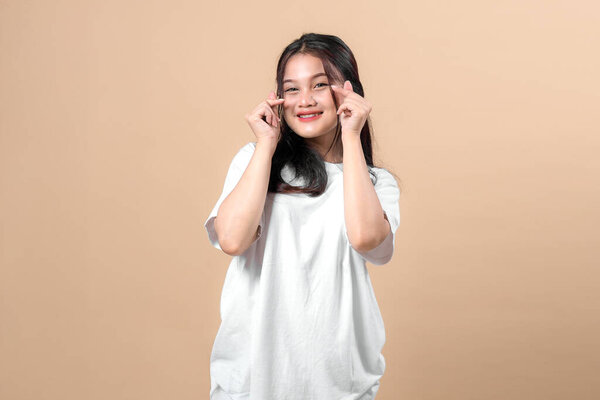 Smiling young Asian woman in a white oversized t-shirt making a double finger heart pose with both hands, looking at the camera confidently against a beige background.