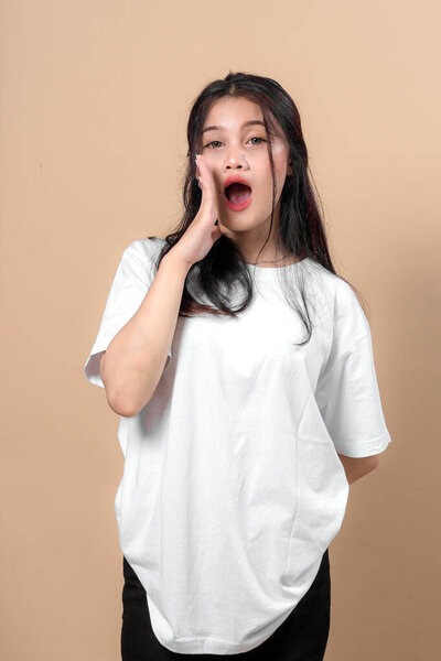 Young Asian woman shouting or calling out with hand near mouth, wearing a white t-shirt and standing against beige background, conveying urgency or spreading important news.