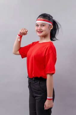 Kırmızı tişörtlü ve siyah pantolonlu enerjik genç kadın, Endonezya 'nın Bağımsızlık Günü' ne sevinç ve vatanseverliğini ifade etmek için kırmızı-beyaz aksesuarlar takarak gururla yumruğunu kaldırıyor..