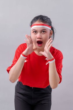 Kendine güvenen Endonezyalı genç kadın, Bağımsızlık Günü 'nü gururla anmak için elleri ağzının kenarında bağırıyor, kırmızı elbiseler ve aksesuarlar giyiyor..