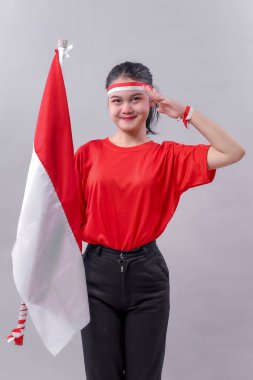 Kırmızı tişörtlü genç Endonezyalı kadın elinde ulusal bayrak tutarken gururla selam veriyor. Bağımsızlık Günü 'nde vatanseverliğini, ruhunu ve sevgisini göstererek özgüvenle gülümsüyor..