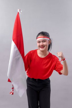 Mutlu bir Bağımsızlık Günü kutlaması için kırmızı ve beyaz giyinmiş, Endonezya bayrağını tutarken gülümseyen ve yumruğunu kaldıran kendinden emin genç bir kadın..