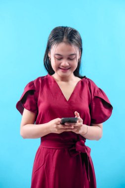Kırmızı elbiseli genç bir Asyalı kadın mavi arka planda akıllı telefonuyla yazarken gülümsüyor, iletişimi, dijital yaşam tarzını, sosyal medyayı ve iş konseptini temsil ediyor..