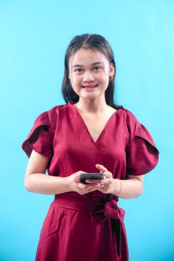Kırmızı elbiseli, gülümseyen genç bir Asyalı kadın mavi arka planda akıllı telefonu tutarken dijital hayatı, iletişimi, iş başarısını, güveni ve modern bağlantıyı sembolize ediyor..