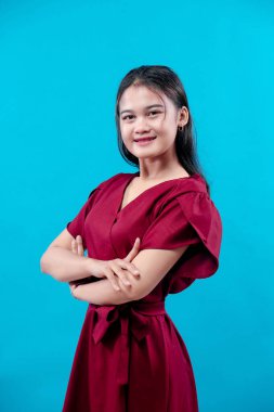 Kırmızı elbiseli, kendine güvenen genç Asyalı kadın kollarını kavuşturup, içten bir şekilde gülümsüyor, zarafet, pozitiflik ve profesyonel cazibesini parlak mavi bir arka plana karşı gösteriyor..