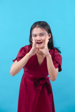 Kırmızı elbiseli genç bir kadın mavi arka planda poz veriyor. Ellerini ağzının yanında tutarken gülümsüyor. Bağırıyor ya da bağırıyor gibi..