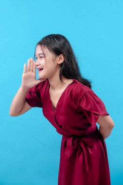 Kırmızı elbiseli genç bir kadın mavi arka planda poz veriyor, elini ağzının yanında kaldırırken gülümsüyor sanki bağırıyor ya da birine sesleniyormuş gibi..