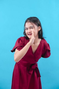 Kırmızı elbiseli neşeli genç bir kadın mavi bir arka plana karşı poz veriyor. Parlak bir gülümsemeyle elini ağzına doğru kaldırıyor..