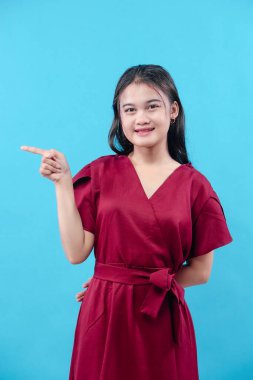 Gülümseyen kırmızı elbiseli genç bir kadın parmaklarını yana doğru gösteriyor, mavi arka planda izole edilmiş, yönü, rehberliği ve pozitif ifadeyi temsil ediyor..