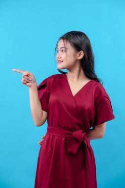 Kırmızı elbiseli genç kadın parmaklarıyla yan tarafı gösterirken kendine güvenen mavi arka planda izole edilmiş rehberlik, yön, odaklanma ve pozitif tutumu sembolize ediyor..