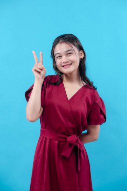 Kırmızı bir elbise giyen neşeli genç bir kadın parmaklarıyla barış işareti gösterirken ışıl ışıl gülümsüyor mavi arka planda güvenle duruyor, neşe ve pozitifliği simgeliyor..
