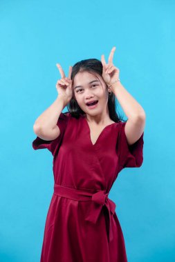 Kırmızı elbiseli neşeli genç bir kadın iki elini kaldırarak poz veriyor, neşeli parmak hareketleri yapıyor, parlak mavi bir arka plana karşı doğal bir güvenle gülümsüyor..