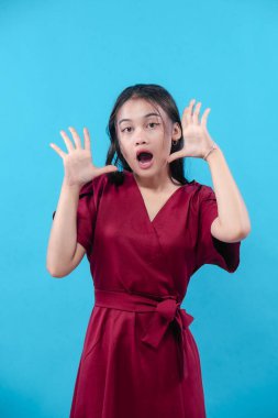 Kırmızı elbiseli genç bir kadın, elleri havada ve ağzı açık bir şekilde şaşkınlığını dile getiriyor. Parlak mavi arka planda dramatik duygular sergiliyor..