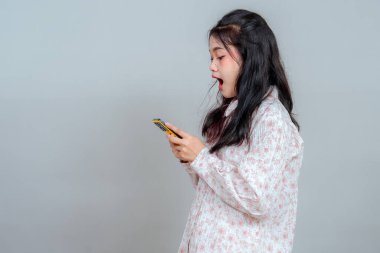 Şaşırmış genç bir kadın akıllı telefonuna geniş gözlü ve ağzı açık bakıyor, şok ya da inançsızlığını ifade ediyor, modern bir stüdyo resminde ham duygu ve dijital tepkiyi yakalıyor..