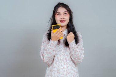 Uzun siyah saçlı genç Asyalı kadın, sarı akıllı telefonu tutarken heyecanını ve umudunu dile getiriyor. Neşeli gözlerle bakıyor ve yumruğunu sımsıkı tutuyor..