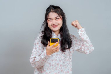 Akıllı telefonu tutarken gülümseyen ve yumruğunu zafer için kaldıran neşeli, genç Asyalı kadın. Pozitif yüz ifadesi, kazanmak için ideal, başarı ve dijital yaşam tarzı kavramları.