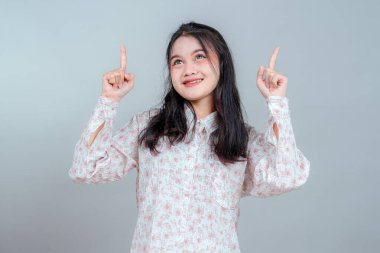Neşeli, genç Asyalı bir kadın her iki işaret parmağıyla işaret ederken gülümseyip yukarı bakıyor. Heyecan, iyimserlik ve canlı bir stüdyo resminde beklenti gösteriyor..