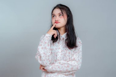 Uzun siyah saçlı, düşünceli bir yüz ifadesi takınan genç Asyalı bir kadın, parmak dudaklarında, düşünceli bir şekilde yukarı bakıyor. Stüdyoda merak ve şüphe dolu bir yüz..