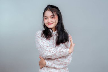 Gülümseyen Asyalı genç bir kadın hafif ama kendine güvenen bir yüz ifadesi sergilerken nazikçe kollarını kavuşturarak dostluk, sakinlik ve stüdyo çekimlerinde sakin bir pozitiflik sergiliyor..