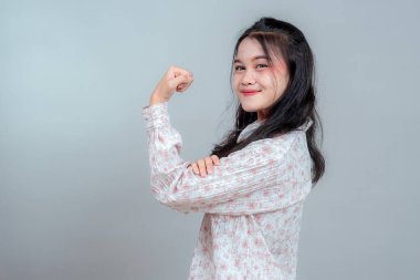 Kendine güvenen, gülümseyen ve pazı gücü gösteren genç Asyalı kadın, güçlendirmeyi, motivasyonu ve pozitif enerjiyi sembolize ediyor. Sade arka planda izole edilmiş.