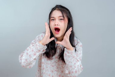 İki eli ağzında bağıran genç Asyalı kadın portresi, aciliyeti ya da heyecanı ifade ediyor. Çiçekli bluz giyiyor, tarafsız gri arka planda poz veriyor..