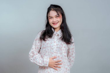 Genç Asyalı kadın elini karnına koyarken parlak bir şekilde gülümsüyor. Onun mutlu ve kendinden emin yüz ifadesi neşe, dostluk ve sıcak, cana yakın bir kişilik taşır..