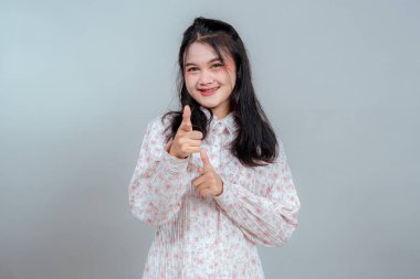 Neşeli genç Asyalı kadın gülümsüyor ve her iki işaret parmağını şakacı, kendinden emin bir jestle işaret ediyor. Reklam ve pazarlama için mükemmel bir yüz ifadesi..