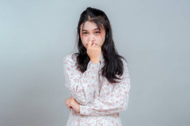 Endişeli, genç bir Asyalı kadının endişeli ve kararsız bir şekilde parmağını ısırmasının yakın plan portresi. Gri arka planda izole edilmiş. Şüphe, gerilim ve duygusal stres kavramı.