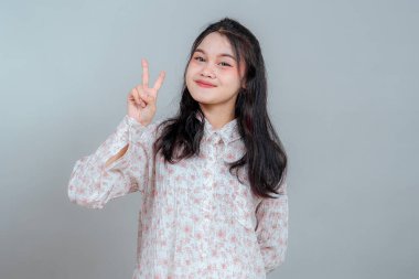 Genç Asyalı kadın parmaklarıyla barış işareti gösterirken özgüvenle gülümsüyor. Çiçekli bir gömlek giyiyor ve neşeli, olumlu bir tavır sergiliyor..