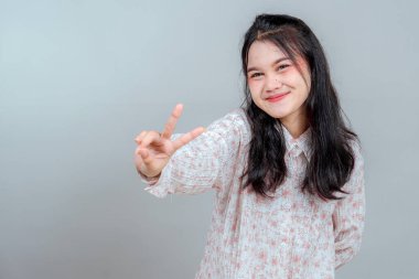 Genç Asyalı kadın kameraya barış işareti tutarken sıcak bir gülümseme sergiliyor. Onun dostane ve neşeli yüz ifadesi mutluluğu ve ulaşılabilirliği yansıtıyor..
