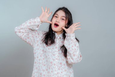 Uzun siyah saçlı, neşeli, genç Asyalı bir kadının portresi. Şaşırmış ve neşeli bir yüz ifadesi takınıyor. Elleri yüzünün yanında, arka planda..