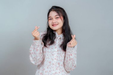 Çift parmaklı kalp hareketi gösterirken, kendine güvenen genç Asyalı bir kadının portresi. Dost canlısı ifade, sade arka planda izole edilmiş, reklam için mükemmel..