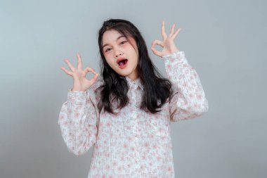 Heyecanlı Asyalı genç bir kadın. İfade dolu bir yüzü var. İki kere el hareketi yapıyor. Ağzı açık gülümsüyor, çiçek desenli bir bluz giyiyor ve heyecanını gösteriyor..