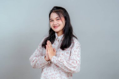 Uzun siyah saçlı, gülümseyen genç Asyalı kadın iki eliyle saygılı bir selamlama hareketi yapıyor. Onun sıcak yüz ifadesi kültürel, konukseverlik ve hoş geldin temalı reklamlara uyuyor..