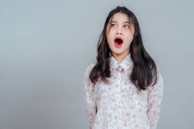 Geniş ağızlı ve şaşırmış bir ifadeyle yukarı bakan genç Asyalı kadın portresi, tarafsız bir arkaplana karşı durarak şaşkınlık, inançsızlık ya da ani bir farkındalık ifade ediyor..