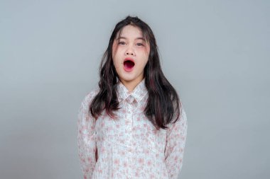 Geniş ağızlı ve şaşırmış bir ifadeyle genç bir Asyalı kadın, düz bir arkaplana karşı duruyor. Şok olmuş yüzü ve vücut duruşu inançsızlığını, korkusunu ya da ani farkındalığını gösteriyor..