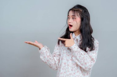 Şaşırmış ve hayrete düşmüş bir yüz ifadesi olan Asyalı genç bir kadın sanki bir ürünü sunar ya da gösteriyormuş gibi avucunun içini işaret ediyor. Gri arka planda izole edilmiş, anlamlı duruş.