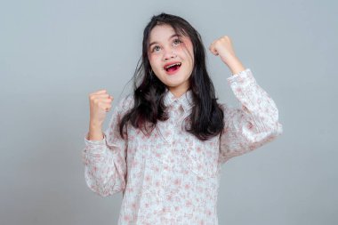 Neşeli bir ifadeyle heyecanlı, genç Asyalı bir kadın. Gözleri açık, ağzı açık, yumruklarını zaferle kaldırıyor. Tarafsız arkaplanda enerjik ve anlamlı bir yüz.