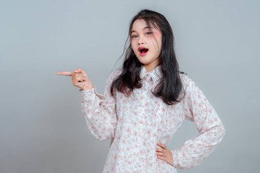 Şaşırmış Asyalı bir kadın, ağzı açık ve şakacı bir ifadeyle işaret parmağıyla işaret ediyor, dikkat ya da yönü gösteriyor. Gri arkaplanda duygusal yüz ile stüdyo çekimi.