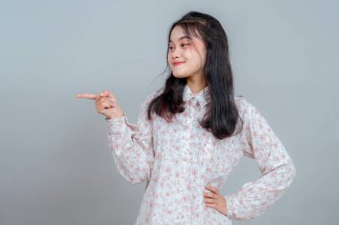 İşaret parmağıyla kenarı işaret ederken tatmin olmuş bir ifadeyle gülümseyen kendinden emin genç Asyalı kadın. Çekicilik, güven ve iletişim konseptini gösteren stüdyo portresi.