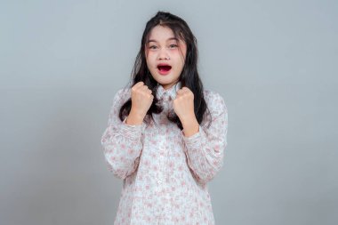 Şaşırmış ve heyecanlı genç Asyalı kadın yumruklarını sıkmış, ağzı açık ve ifade edici bir yüzü var. Duygusal tepki, coşku ve beklenmedik neşeyi gösteren bir stüdyo portresi..