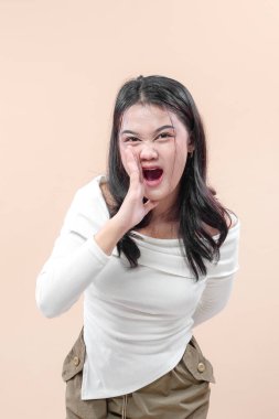 Sıradan kıyafetli, dışavurumcu bir Asyalı kadın eli ağıza yakın öne eğiliyor, bağırıyor ya da enerjik bir şekilde bağırıyor, cesur bir duygu sergiliyor..