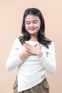 Genç bir Asyalı kadın gözlerini sakin bir gülümseme ile kapatıyor ve elleri göğsünde, minnettarlığı, farkındalığı ve olumlu duygusal dengeyi simgeliyor..