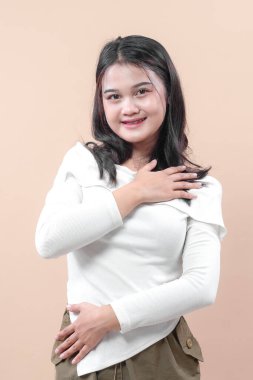 Gülümseyen Asyalı bir kadın bir elini göğsüne diğerini beline koyar, sıcaklığını, minnettarlığını ve kendine olan güvenini olumlu yaşam tarzına yansıtır..