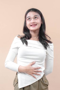 Asyalı beyaz bluzlu bir kadın elini karnına koyup yukarı bakarken nazikçe gülümser. Mutluluk, minnettarlık ve olumlu duyguları ifade eder..