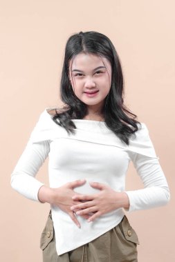 Yumuşak bir gülümsemesi olan Asyalı bir kadın karnına dokunuyor, sıcaklığını ve pozitifliğini ifade ediyor, sağlık, yaşam tarzı ve refah kavramları için mükemmel..