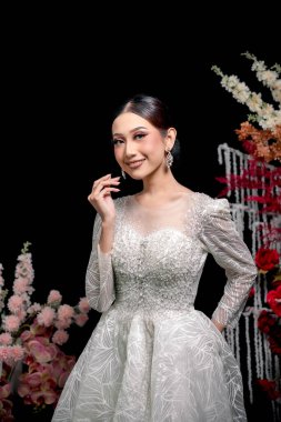 Çekici Endonezyalı gelinlik içinde kristal boncuklu detaylar, ışıl ışıl makyaj ve parlak küpeler, canlı renklerle çiçeklere karşı zarif bir şekilde gülümseyen.